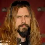 Rob Zombie with long hair and sunglasses, posing for a portrait against a red backdrop.
