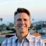 Portrait of a man with short hair wearing a checkered shirt, smiling at the camera outdoors.